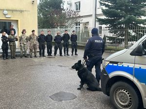 Grupa młodzieży w mundurach wojskowych stojąca przy budynku przed nimi stoi umundurowany policjant z psem służbowym obok radiowozu.