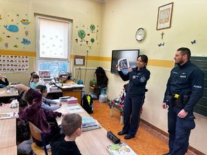 Policjantka stojąca w szkolnej klasie obok niej umundurowany policjant. Policjantka trzyma w ręce wydrukowany znak drogowy i pokazująca go dzieciom siedzącym w szkolnych ławkach.
