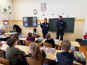 Policjantka stojąca w szkolnej klasie obok niej umundurowany policjant. Policjantka trzyma w ręce policyjną tonfę  i pokazująca go dzieciom siedzącym w szkolnych ławkach.