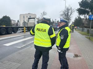 Dwóch policjantów stojących na chodniku przy jezdni z oznakowanym przejściem dla pieszych
