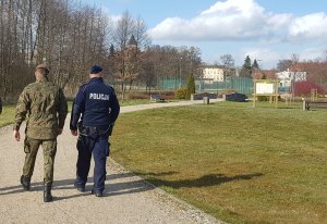 Policjsnt oraz żołnierz wot iddą aleją w parku w tle widać w oddali budynki.