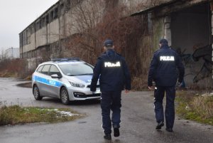 Policjanci na tle budynku do którego sie zblizają. W tle stoi radiowóz.