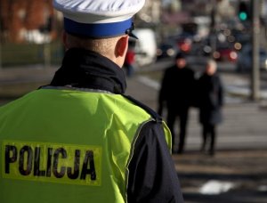 Policjant stoi przy przejściu dla pieszych w dali widać dwie osoby przechodzące przez przejście