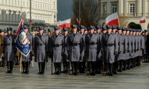 Policjanci stoją w kolumnie w płaszczach galowych. W pierwszym rzędzie policjant trzyma sztandar. W tke widać powiewajace flagi