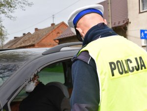 Policjant stoi przy kontrolowanym pojeździe- widać plecy policjanta, który jest ubrany w kamizelke z napisem policja