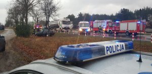 Na pierwszyym planie widać oświetlenie na dach radiowozu z napisem policja- w tle miejsce zdarzenia drogowego- rozbite auto osobowe w rowie, na jezdni samochód ciężarowy oraz karetke pogotowia i samochody straży pożarnej