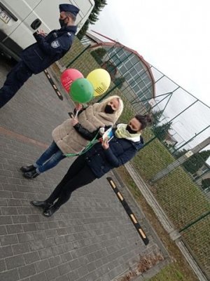 Policjant sprawdza dane osobowe stojąc przy pojeździe- bliżej stoja dwie kobiety z balonami w ręku