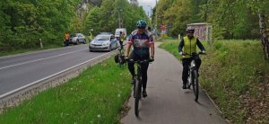 Dwóch rowerzystów na pierwszym planie jedzie ścieżka rowerową a z tyłu po ulicy jedzie za nimi radiowóz