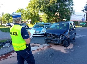 Policjantka prowadzi oględziny rozbitego auta. W tle policyjny radiowóz, a obok rozbity przód granatowego auta osobowego.