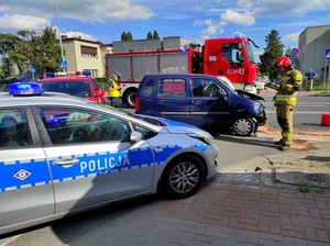 Na pierwszym planie prawy bok radiowozu policyjnego. Tuż za nim policjant i strażak pracują na miejscu zdarzenia drogowego.