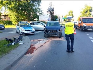 Policjantka prowadzi oględziny rozbitego auta. W tle policyjny radiowóz, a obok rozbity przód granatowego auta osobowego.