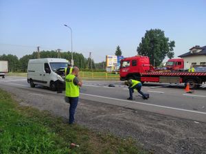 policjant stojący na poboczu, za nim rozbity samochód i policjant robiący pomiary