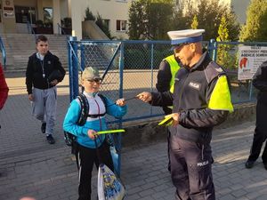 POLICJANT STOJĄCY PRZED SZKOŁĄ WRĘCZ CHŁOPCU OPASKĘ ODBLASKOWĄ