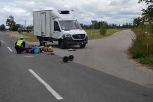 rozbity samochód stojący na poboczu, na drodze leży przewrócony motorower obok pracujący policjanci