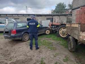 umundurowany policjant na tle budynków gospodarczych oraz samochodu osobowego i traktora z przyczepką