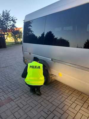 policjant w żółtej kamizelce odblaskowej z napisem POLICJA schylający się prze tylnym kole szarego busa