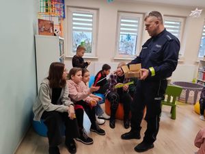 policjant rozdający elementy odblaskowe w bibliotece wokół siedzi grupka dzieci