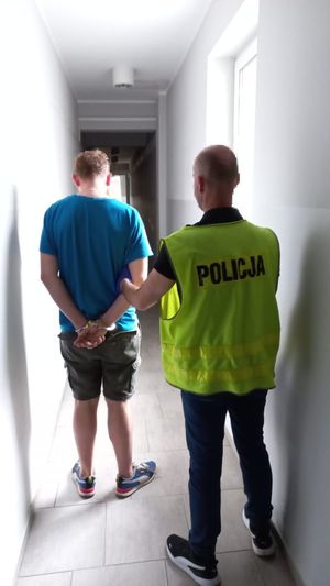 zatrzymany mężczyzna w niebieskiej koszulce prowadzony przez policjanta w żółtej kamizelce