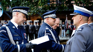 Umundurowani Policjanci w mundurach galowych wręczający nominacje umundurowanemu policjantowi w mundurze galowym z białą czapką.