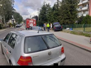 Stojący srebrny samochód, za nim na chodniku stoi inny czarny samochód, przy którym stoją policjanci i strażacy.