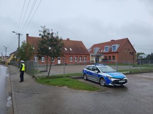 Policyjny radiowóz stojący na parkingu przed budynkiem szkoły na chodniku obok stoi policjant w żółtej kamizelce odblaskowej