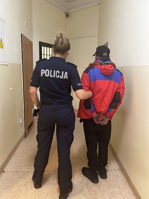 Skuty w kajdanki mężczyzna prowadzony przez umundurowaną policjantkę.