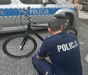 Policjant schylający się przy rowerze stojącym na tle rawiowozu.