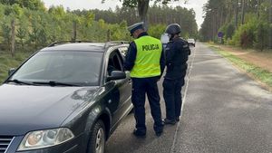 Dwóch policjantów stojących na jezdni przy ciemnym samochodzie  rozmawiających z jego kierowcą