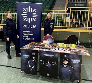 Dwie umundurowane policjantki stojące przy banerze KPP w Chojnicach przed nimi stół z artykułami promującymi pracę w policji