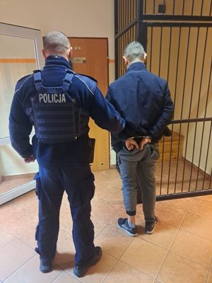 Mężczyzna skuty kajdankami stojący tyłek=m obok niego stoi policjant, który trzyma go za nadgarstki