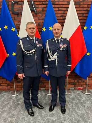 Dwóch policjantów w mundurach galowych stojących na tle flag.