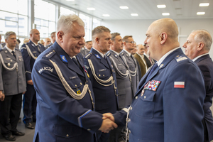 Policjant w umundurowaniu galowym policji  ściskający dłoń innemu umundurowanemu policjantowi stojącemu w szeregu z innymi policjantami