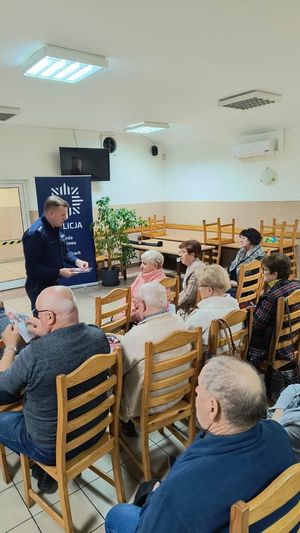 Policjant w sali stojący przed grupą starszych osób siedzących na krzesłach.