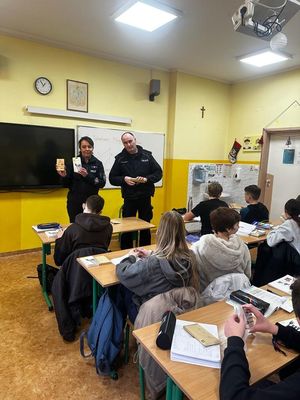 Policjantka i policjant stojący w szkolnej klasie przy tablicy rozmawiający z uczniami siedzącymi w szkolnych ławkach.