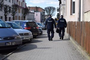 Dwóch policjantów idących chodnikiem, przy którym stoją zaparkowane samochody.