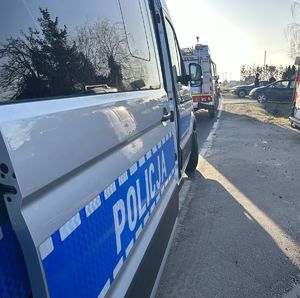 Napis Policja widoczny na otwartych drzwiach radiowozu stojącego na miejscu zdarzenia drogowego. Za radiowozem stoi pojazd staży pożarnej