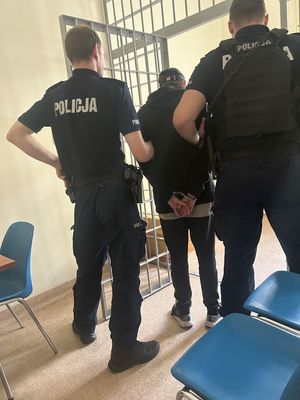 Dwóch policjantów na tle kraty trzymających zatrzymanego