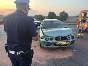 Policjant stojący na jezdni obok rozbitego pojazdu koloru zielonego sprawdzający jego uszkodzenia. Obok stoi wóz strażacki a przy nim strażak w tle widoczny zachód słońca