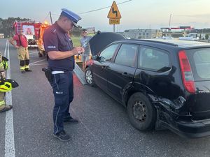 Policjant stojący na jezdni, który spisuje uszkodzenia czarnego samochodu osobowego, który brał udział w zdarzeniu drogowym. Za min stoi wóz strażacki oraz strażak