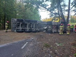 Na jezdni leży wywrócona ciężarówka, z której naczepy wysypał się piasek. Obok chodzą strażacy oraz policjant