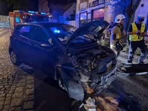 w nocy ciemny samochód z rozbitym przodem stojący na jezdni obok policjant i strażak