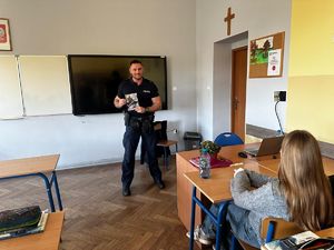 Policjant stojący w szkolnej klasie przed uczniami, którzy siedzą w ławkach, trzymający ulotkę zachęcającą do wstąpienia do policji.