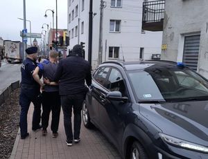 Policjant w mundurze oraz policjant w ubraniu cywilnym prowadzący osobę zatrzymaną chodnikiem.