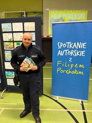 Policjant na hali sportowej podczas pogadanki z dziećmi stojący przy niebieskim banerze z książką w ręku