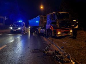 W nocy na środku mokrej jezdi stoi policyjny radiowóz obok niego ciężarówka przed nią lezy przewrócony rower ze śladami po zdarzeniu drogowym.