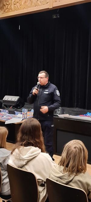 Policjant w sali stojący przed grupą osób siedządzych prze nim.