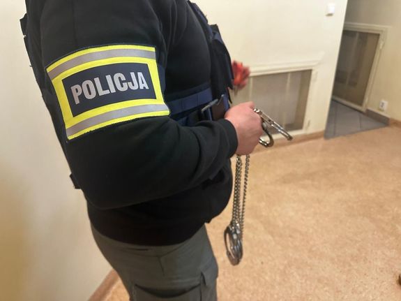 Nieumundurowany policjant z opaską policja na ręku trzymający kajdanki w dłoni.