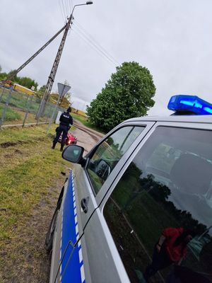 Policajantka stojąca przed radiowozem obok czerwonego motocykla któy leży na trawie obok ogrodzenia.