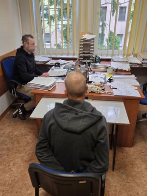 Policjant siedzący ptrzy biurku przesłuchujący zatrzymanego który siedzi obok niego