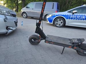 Hulajnoga elektryczna stojąca obok samochodu z uszkodzonym przednim zdarzakiem, prze dnimi w tle policyjny radiowóz.